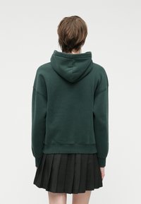 Dark green hoodie with a relaxed fit, large hood, and ribbed hem. Styled over a black pleated skirt, showcasing contrasting textures.