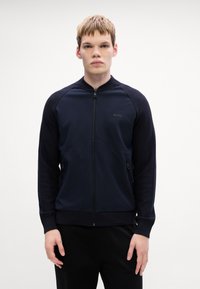 Mornarska atletska zip majica z kontrastnimi pletenimi rokavi, ki ima logo na prsih in dva stranska zip žepa. Gladka tekstura.