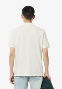 Witte polo met korte mouwen, zachte textuur en een klassieke kraag, gecombineerd met lichtblauwe jeans en een groene trui in de hand.