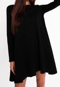 Femme aux longs cheveux bruns portant une robe noire ample à manches longues avec un ourlet asymétrique, posant devant un fond blanc.