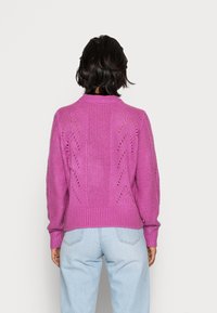 Stickad tröja i klar rosa, med ribbad krage och ärmslut, öppenhetsdesign på baksidan och distinkt texturerat mönster.