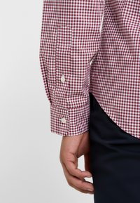 Handen vilar vid sidan och bär en långärmad skjorta i röd och vit rutigt mönster med knäppta ärmslut och mörka byxor.