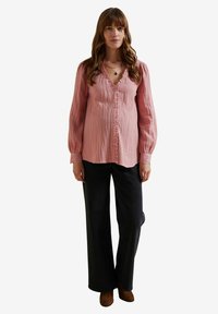 Hellrosa, strukturierte Bluse mit Knopfleiste und Rüschensaum. Mit Ballonärmeln und Spitzenbesatz. Kombiniert mit dunklen, weiten Jeans.