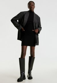 Zwart geribbeld jurkje onder een oversized grijs geruit blazer, gecombineerd met hoge zwarte leren laarzen met dikke zolen. Eenvoudige achtergrond.