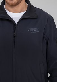 Schwarze Zip-Jacke mit hohem Kragen, ausgestattet mit einem dezenten "BOMBERS ORIGINAL" Logo und glattem, strukturiertem Stoff.