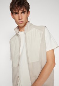 Lys beige ermeløs vest med glidelås foran, med en blanding av teksturert fleece og glatte stoffpaneler. Enkelt design, minimale detaljer.