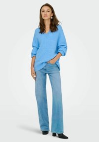 Jasnoniebieski, oversized sweter z dekoltem w kształcie V i mankietami, zestawiony z jasnoniebieskimi dżinsami w fasonie dzwonów oraz czarnymi botkami. Minimalistyczny design.