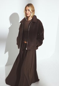 Una donna bionda indossa una giacca a zip texture marrone scuro sopra un top marrone e una gonna lunga e fluente, in piedi davanti a uno sfondo bianco.