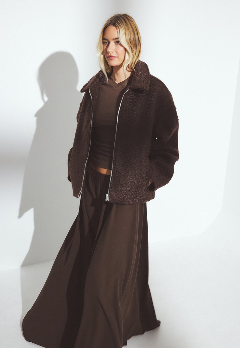 Una donna bionda indossa una giacca a zip texture marrone scuro sopra un top marrone e una gonna lunga e fluente, in piedi davanti a uno sfondo bianco.