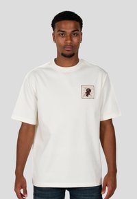 Krémové bavlněné tričko s krátkým rukávem, s čtvercovou kapsou a grafickou ilustrací v hnědých tónech na levé části hrudi.