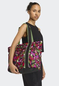 Mujer con cabello rizado y top negro sin mangas sosteniendo un bolso grande con estampado de aves coloridas y asas con rayas verdes.