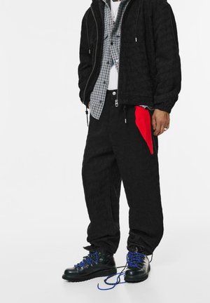 Veste matelassée noire avec capuche, chemise à carreaux grise en dessous, pantalon noir avec une poche d'accent rouge et bottes noires avec des lacets bleus.