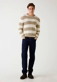 Pull beige à rayures blanches avec texture côtelée, associé à un jean bleu foncé. Le mannequin se tient sur un fond clair, portant des chaussures sombres.
