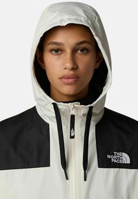 Biała i czarna kurtka z kapturem na zamek błyskawiczny, z logo na piersi oraz kontrastującymi panelami na ramionach. Teksturowany materiał i sznurki do regulacji.