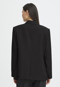 Blazer noir sur mesure avec col cranté, épaules structurées et texture lisse, doté d'une seule couture arrière et de manches longues.