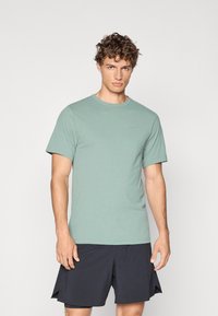 Camiseta verde de cuello redondo hecha de una tela suave, con mangas cortas y un logotipo sutil. Llevada con pantalones cortos deportivos negros.