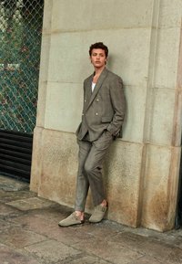 Šedý kostkovaný dvouřadý oblek s dvěma knoflíky, slim fit kalhoty a taupe dřeváky. Hladká látka s jemným leskem, stojící u zdi.