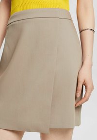 Gonna beige con una silhouette ad A pulita, caratterizzata da una texture liscia e un delicato dettaglio a pieghe, abbinata a un top a coste giallo.