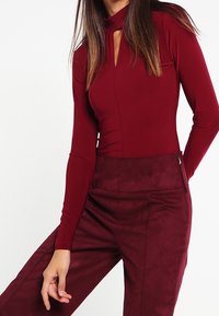 Burgundy långärmad bodysuit med nyckelhålskrage, kombinerad med matchande högmidjade texturerade byxor. Slätt, tight material genom hela.