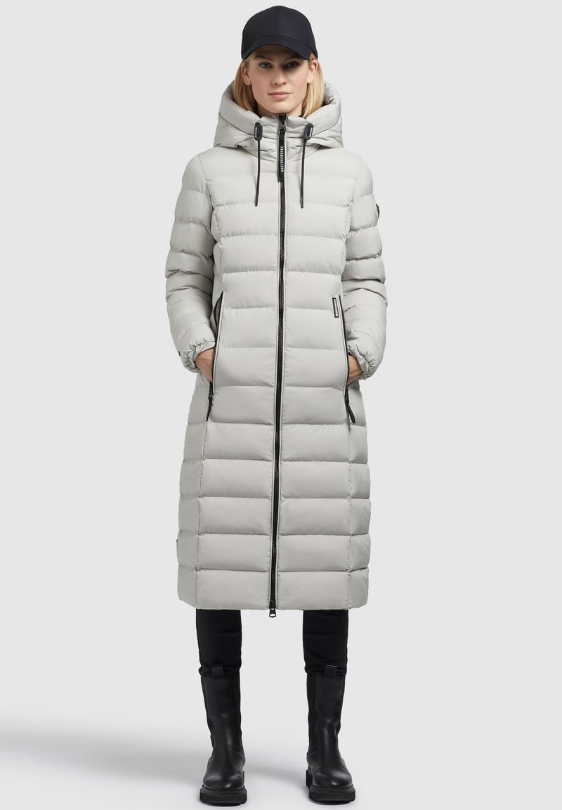 Hellgraue Daunenjacke mit Kapuze, vertikalen Steppungen, einem Reißverschluss vorne und Seitentaschen, kombiniert mit schwarzen Hosen und Stiefeln.