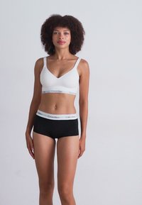 Hvit bralette med V-hals og sorte boyshorts, med en Calvin Klein-logo på livlinningen, vist mot en nøytral bakgrunn.