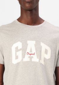 Mand iført en lysegrå t-shirt med store hvide blokbogstaver, der staver "GAP" og en lille rød skrifttype "Original" i midten.