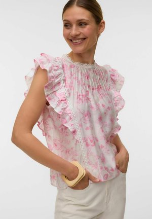 Donna sorridente in camicetta bianca trasparente con stampa floreale rosa, maniche arricciate, bordo in pizzo, pantaloni bianchi e un braccialetto beige spesso.