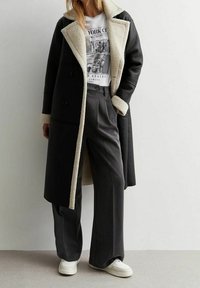 Kvindelig iført lang sort pelstørret coat over en hvid New York City grafisk T-shirt, grå brede bukser og hvide sneakers.