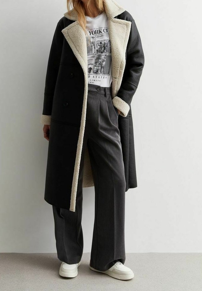 Kvindelig iført lang sort pelstørret coat over en hvid New York City grafisk T-shirt, grå brede bukser og hvide sneakers.