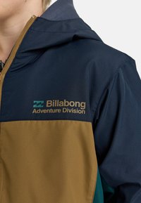 Giacca Billabong con un design a blocchi di colore blu navy e beige. Presenta una zip, cappuccio attaccato e logo "Adventure Division" in oro.