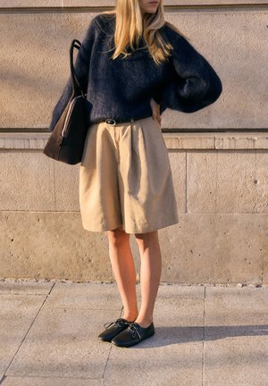 Woman standing outdoors wearing a dark fuzzy sweater, beige knee-length shorts, black lace-up shoes, and carrying a dark brown shoulder bag.