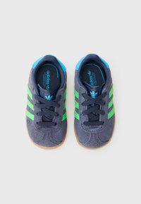 Αθλητικά παπούτσια Adidas με επάνω μέρος από σουέντ σε σκούρο γκρι, πράσινες ρίγες και μπλε λεπτομέρειες. Διαθέτουν μαύρα κορδόνια και σόλα από καουτσούκ χρώματος καραμέλας.