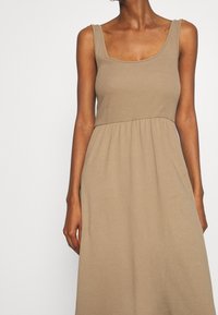 Žena v bez ramínek béžových šatech s přiléhavým žebrovaným vrškem a volnou sukní, zobrazená od krku po střed stehna na jednobarevném pozadí.