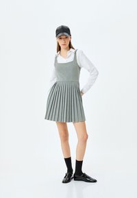 Robe grise plissée avec un décolleté carré et des bretelles, associée à une chemise blanche à boutons, une casquette noire et des chaussures vernies noires.