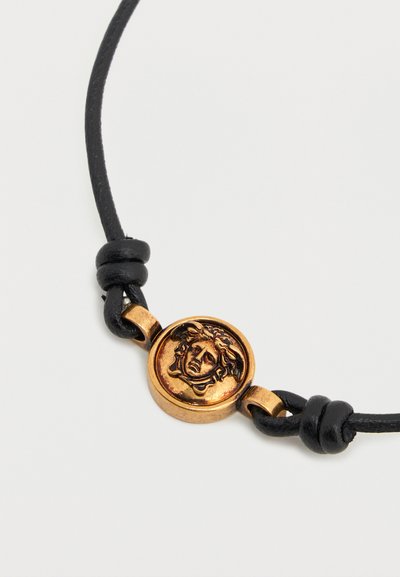 Βραχιόλι με μαύρο κορδόνι και στρογγυλή χρυσή μενταγιόν που απεικονίζει μια λεπτομερή κεφαλή Μέδουσας στο κέντρο.