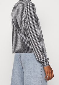 Žena oblečená v dlouhém rukávu blůzu s černobílým vzorem houndstooth, která je v kombinaci se světle modrými džíny s vysokým pasem.