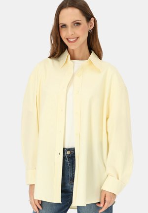 Kobieta ubrana w luźną, jasnżółtą koszulę na guziki na białym topie i niebieskich dżinsach, uśmiechająca się na jednolitym tle.
