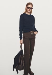 Granatowy sweter, brązowe wysokie spodnie z okrągłą klamrą paska, brązowa torebka z przewieszoną przez nią bluzą oraz ciemne okulary przeciwsłoneczne.