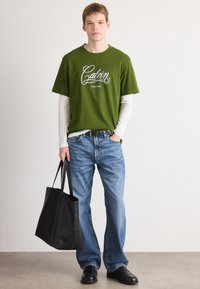 Young man wearing a green Calvin Klein T-shirt over a white long sleeve shirt, blue jeans, black shoes, and holding a large black tote bag.