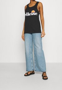 Person, der ein schwarzes ärmelloses Ellesse-Top, hellblaue weit geschnittene Jeans und schwarze Sandalen trägt und vor einem einfarbigen Hintergrund steht.