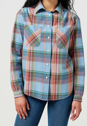 Kobieta w kolorowej, kraciastej koszuli zapinanej na guziki z dwoma kieszeniami na piersiach oraz w niebieskich dżinsach, stojąca z rękami swobodnie opuszczonymi wzdłuż ciała.