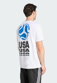 Bijela majica s kratkim rukavima s plavim grafičkim logotipom i ponovljenim tekstom "USA" u podebljanom crnom fontu na leđima. Glatka pamučna tekstura.