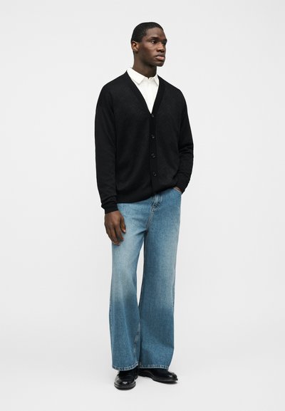 Άνδρας στέκεται με το ένα χέρι στην τσέπη, φοράει μαύρη ζακέτα με κουμπιά, λευκό πουκάμισο με γιακά, μπλε τζιν με φαρδιά μπατζάκια και μαύρα παπούτσια, σε λευκό φόντο.