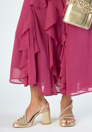 Dobladillo con volantes de vestido magenta con sandalias de tacón grueso doradas y bolso a juego dorado con decoración de joyas.