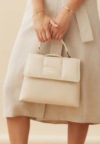 Un sac à main en cuir crème avec une forme structurée, un rabat à l'avant et des détails en métal doré, tenu par des mains au-dessus d'un vêtement de couleur claire.