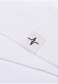Witte ribstof met een gestikt lichtbeige label met een zwart logo op de zoom. De textuur is glad en rekbaar.