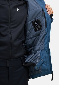 Blau-schwarze Jacke mit gesteppter Innenfütterung, ausgestattet mit einer Reißverschlusstasche, Logo auf der Brust und verstellbaren Bündchen. Sichtbare Etiketten im Inneren.