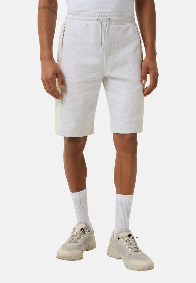 Lichtgrijze katoenen shorts met een trekkoord in de taille, zijritsen en contrasterende crèmekleurige panelen. Draagt witte sokken en beige sneakers.