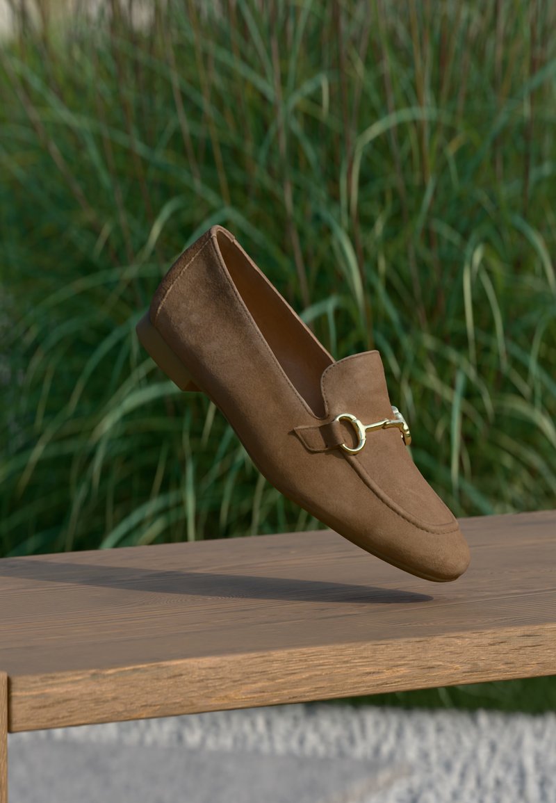 Mocassin en daim marron avec détail mors en or, suspendu au-dessus d'un banc en bois, avec de l'herbe verte en arrière-plan.