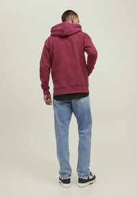 Felpa con cappuccio bordeaux dal taglio comodo, con polsini a costine e cappuccio regolabile, abbinata a jeans azzurri chiari e sneakers scure.